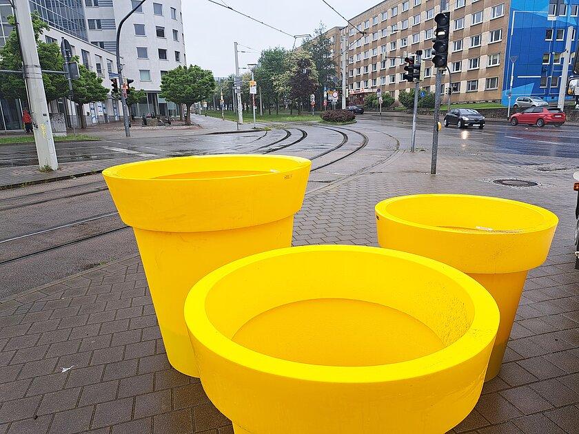 Drei unterschiedlich große, gelbe Pflanzkübel, die an einer Straßenecke stehen