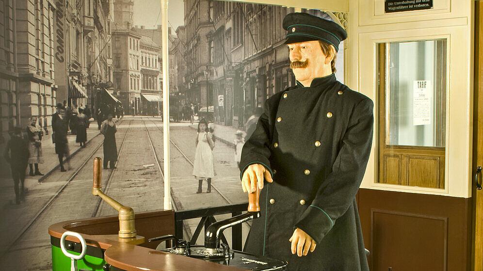 Ausstellungsinszenierung mit einer Straßenbahn um 1900 und Straßenbahnfahrer. Im Hintergrund ist die Schlossstraße mit Straßenbahnschienen zu sehen und eine Werbetafel mit verschiedenen Anzeigen. © Ulrich Fischer