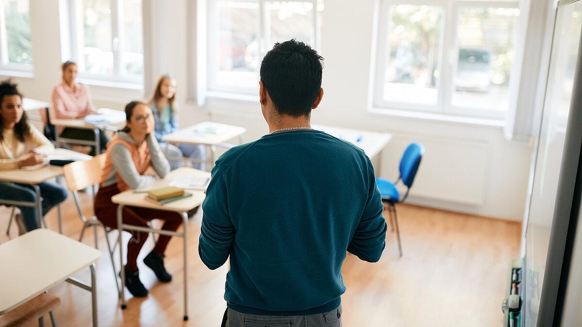 Symbolbild Schule, Schüler sitzen an ihre Tischen, eine Person steht vor der Klasse. © stock.adobe.com
