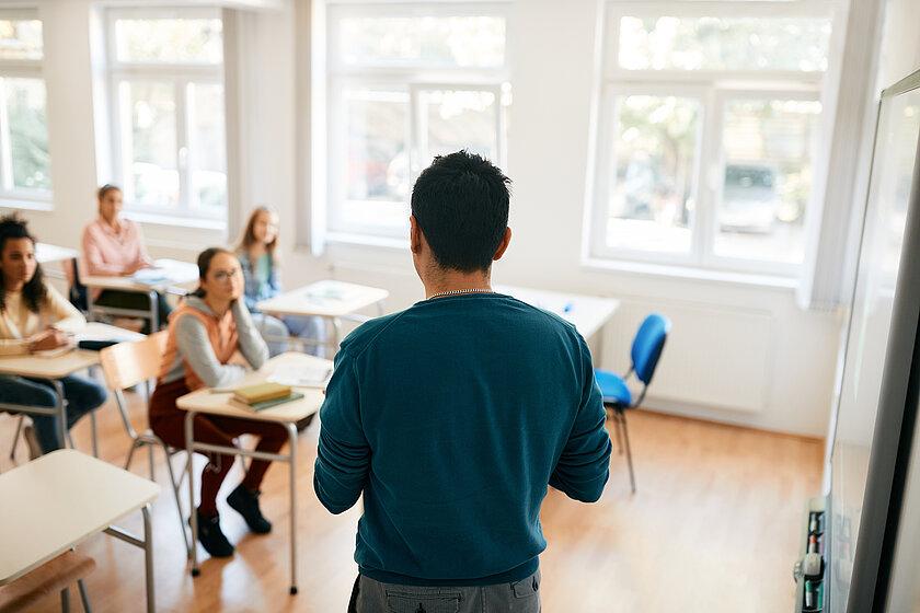 Symbolbild Schule, Schüler sitzen an ihre Tischen, eine Person steht vor der Klasse. © stock.adobe.com