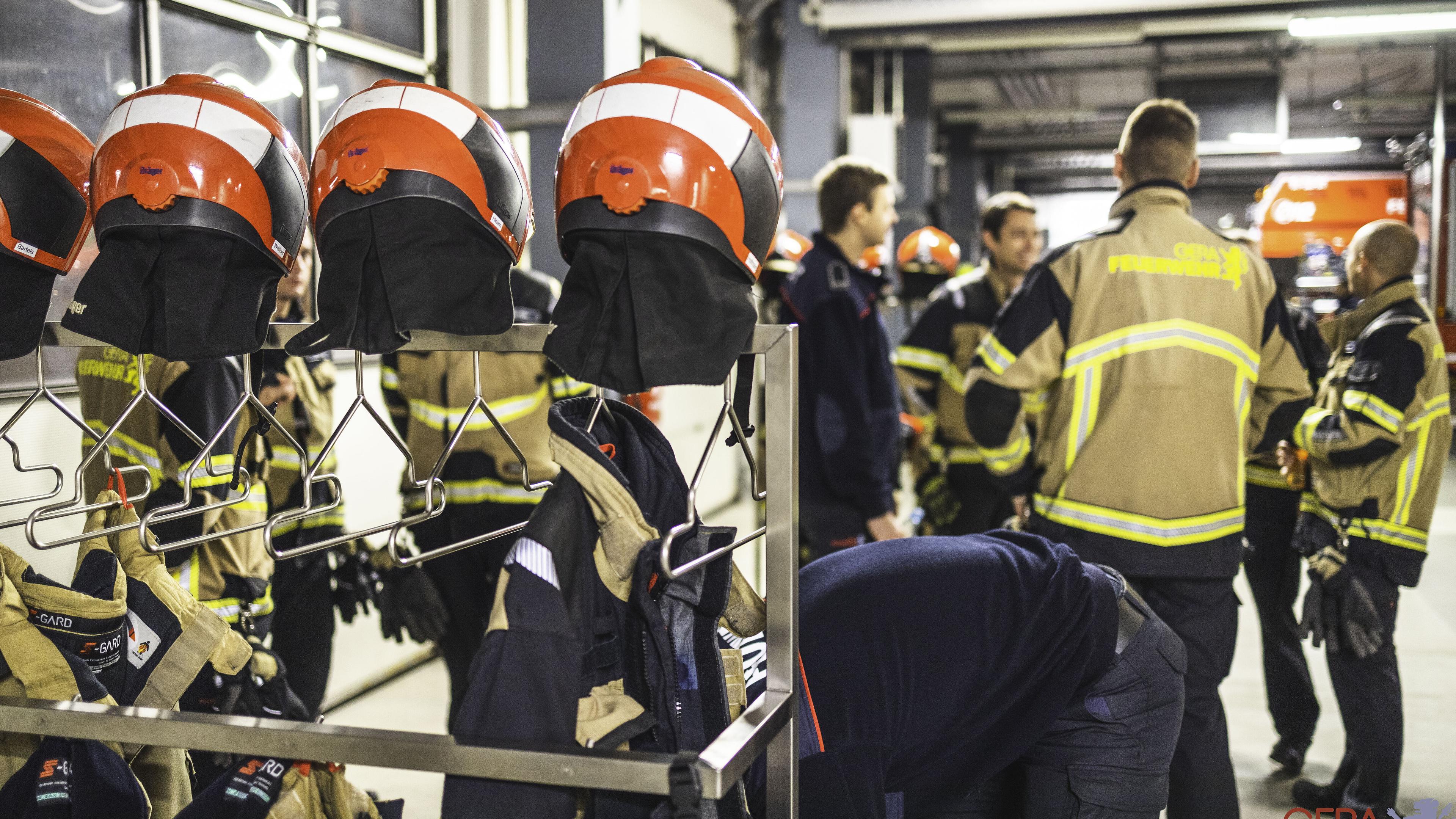 Im Vordergrund hängen an einem Ausrüstungsständer mehrere Feuerwehrhelme und -uniformen, im Hintergrund steht eine Gruppe Feuerwehrleute.