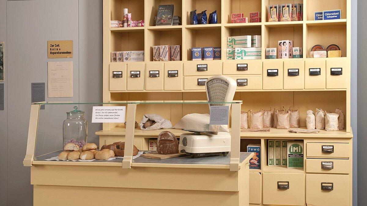 Blick in die Ausstellung - HO-Geschäft um 1950 mit einer Theke auf der sich ein Waage und verschiedene Backwaren befinden. Im Hintergrund sind verschiedene Produkte des täglichen Bedarfs zu sehen, u.a. auch Schubkästen in denen sich Lebensmittel befanden. © Ulrich Fischer