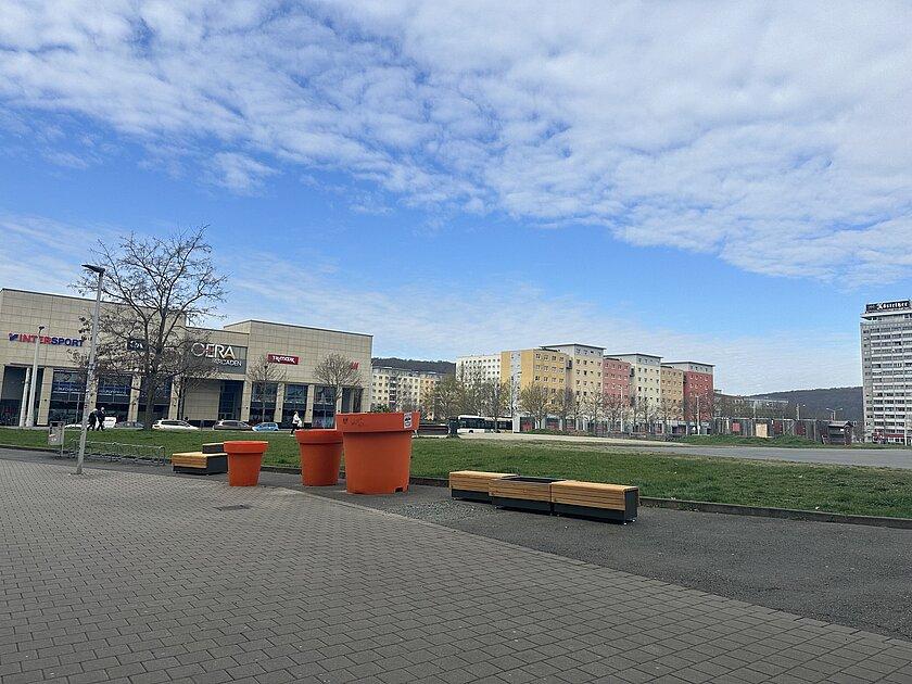 Zwei Hölzerne Bänke vor einer grünen Freifläche, in der Mitte drei orange Planzkübel. © Stadt Gera