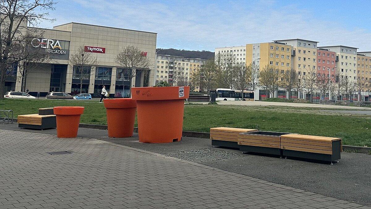 Zwei Hölzerne Bänke vor einer grünen Freifläche, in der Mitte drei orange Planzkübel. © Stadt Gera