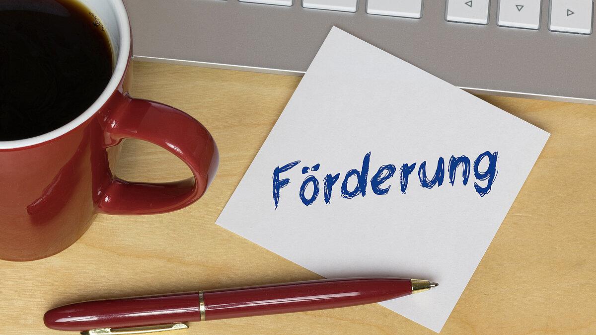 Büroplatz mit einem Zettel auf dem Förderung steht . © Adobe Stock