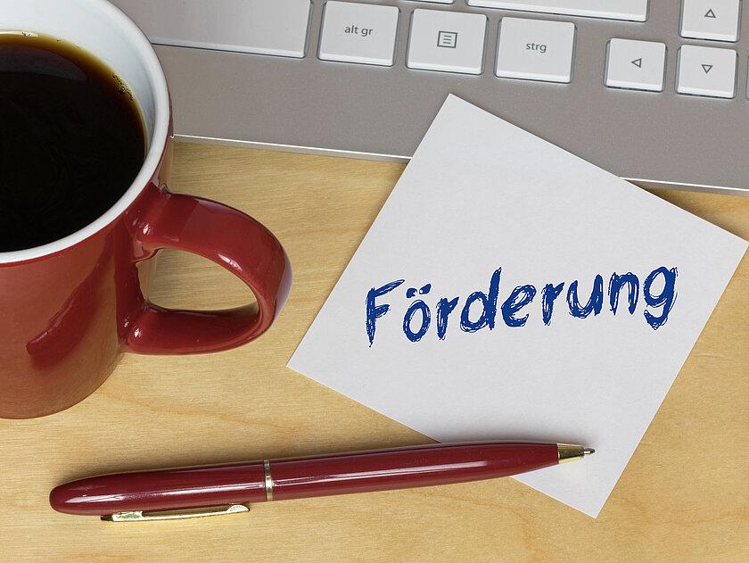 Büroplatz mit einem Zettel auf dem Förderung steht . © Adobe Stock