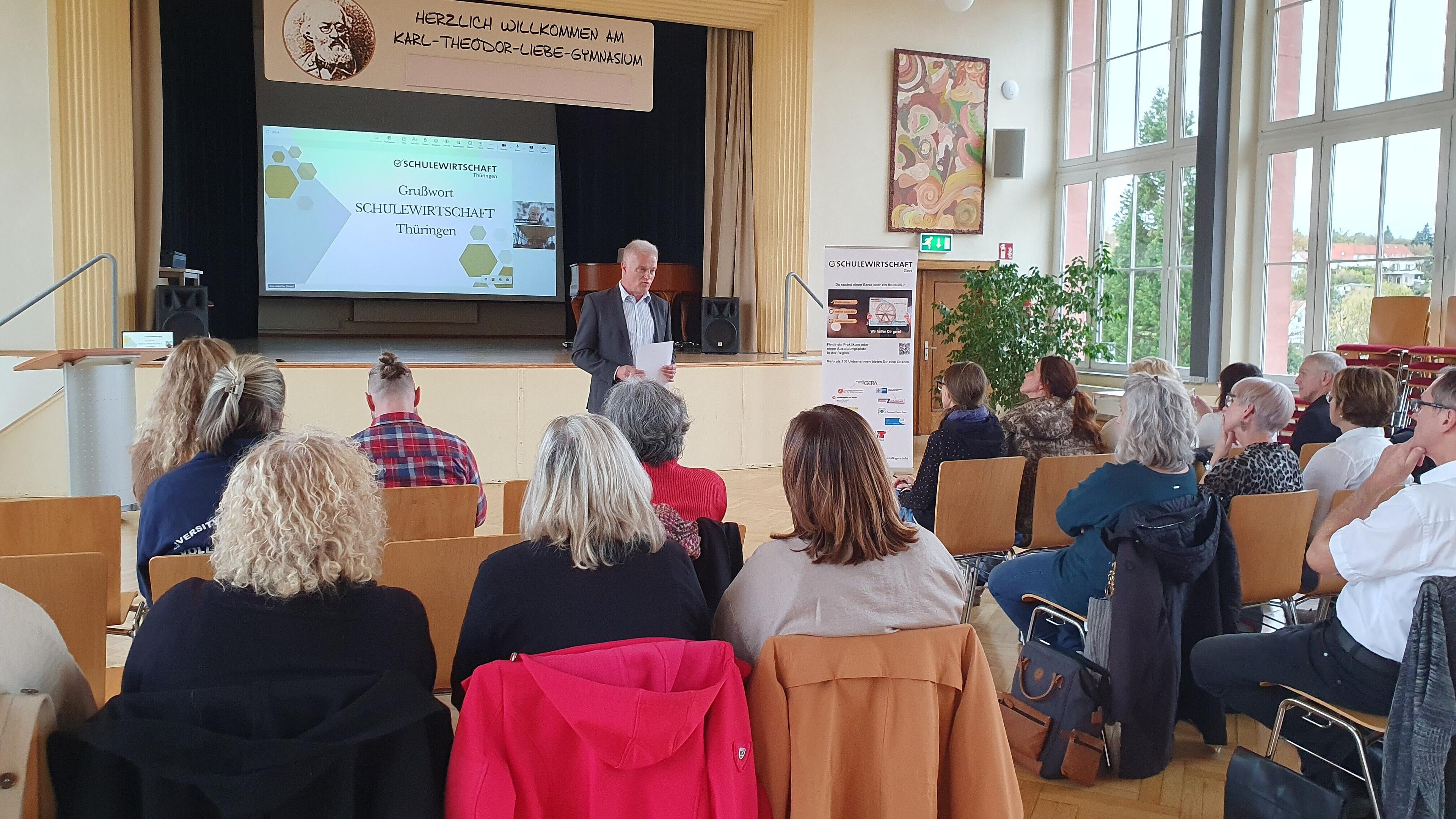 Ein Mann steht in einer Aula vorn und spricht vor einem sitzenden Publikum, welches man von hinten sieht. © IHK Ostthüringen