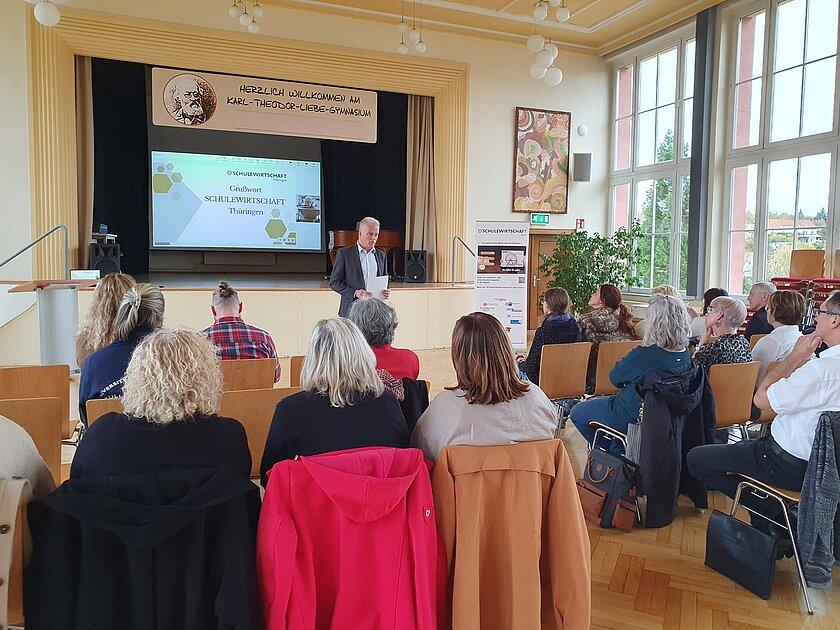 Ein Mann steht in einer Aula vorn und spricht vor einem sitzenden Publikum, welches man von hinten sieht. © IHK Ostthüringen