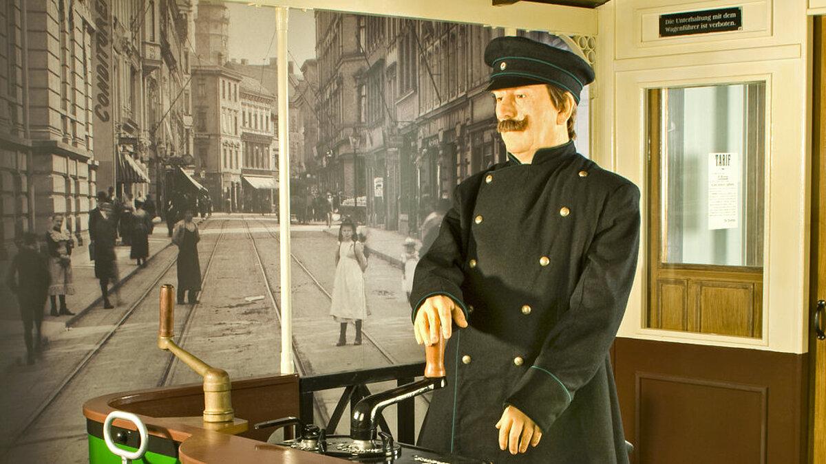 Ausstellungsinszenierung mit einer Straßenbahn um 1900 und Straßenbahnfahrer. Im Hintergrund ist die Schlossstraße mit Straßenbahnschienen zu sehen und eine Werbetafel mit verschiedenen Anzeigen