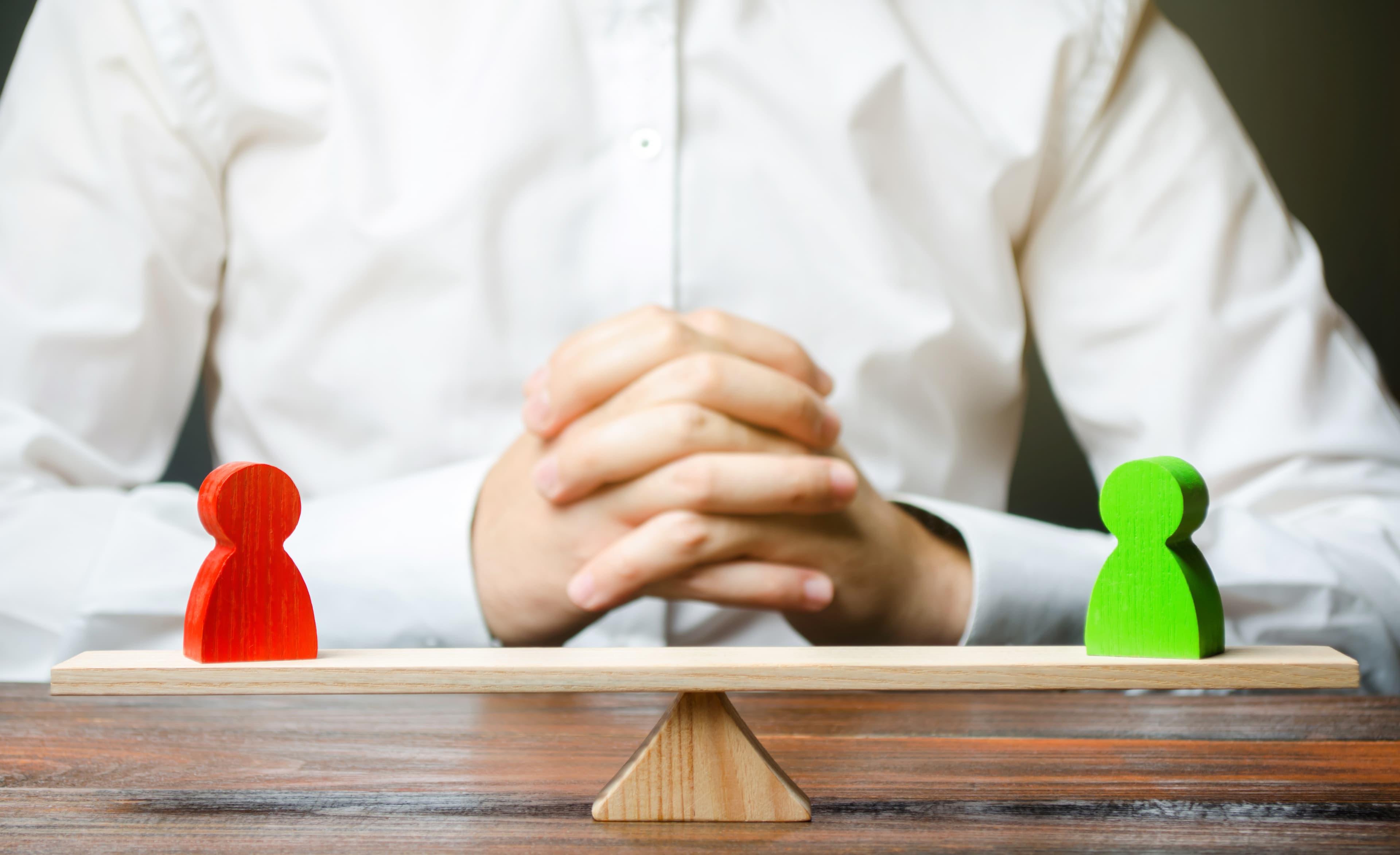 Verbildlichung eines Interessenkonflikts mit einer roten Spielfigur auf der linken und einer grünen Spielfigur auf der rechten Seite einer Holzwaage. Mittig dahinter sitzt eine Person mit gefalteten Händen, die schlichtet.. © Andrii Yalanskyi - stock.adobe.com