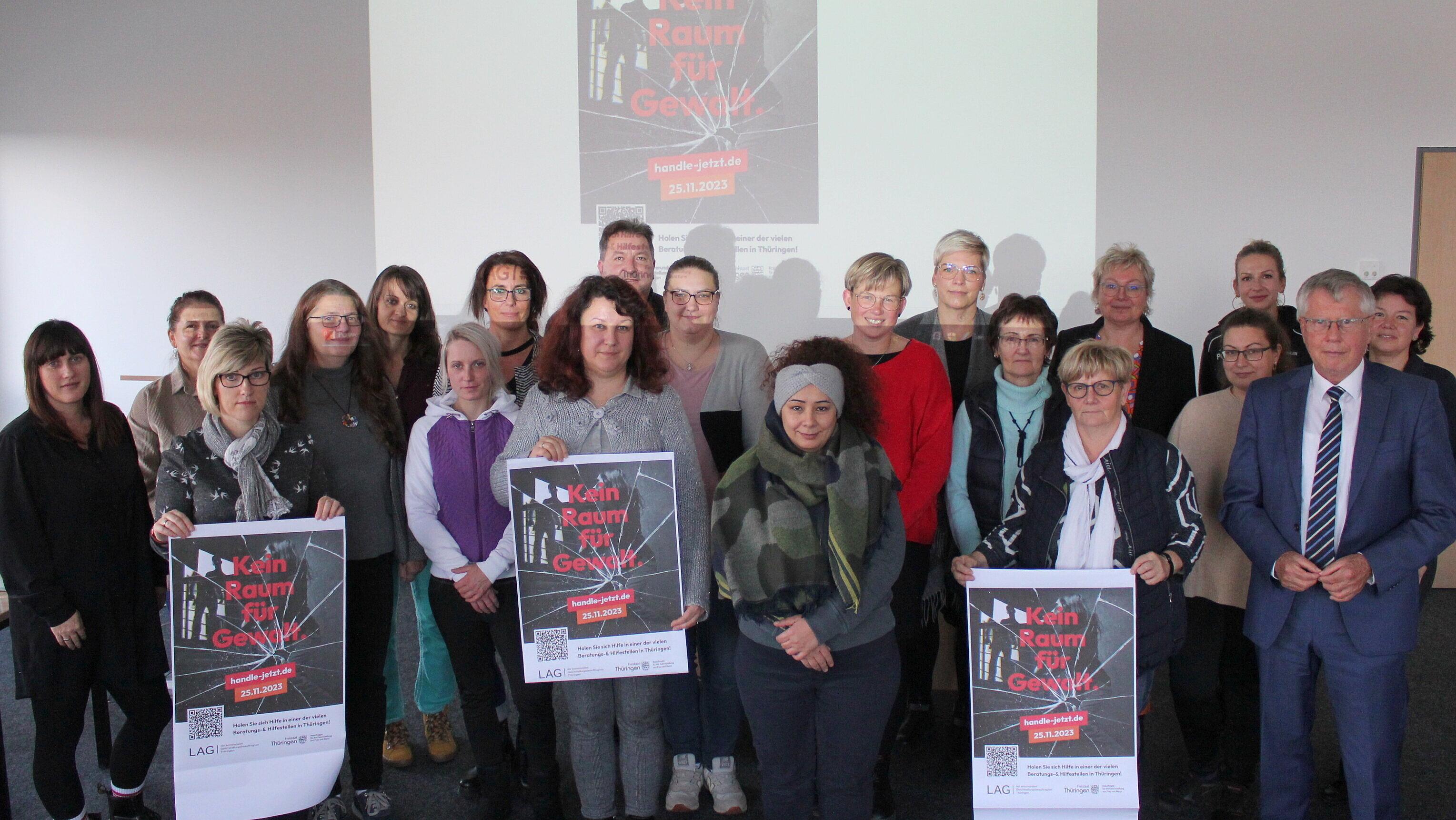 Etwa 20 Personen stehen vor einer Leinwand-Projektion, auf der ein Plakat der Kampagne "handle jetzt" zusehen ist. Die Personen stehen neben- und hintereinander in mehreren Reihen. Drei Frauen in der ersten Reihe zeigen A2-große Plakate zu handle jetzt.. © Saale-Holzland-Kreis