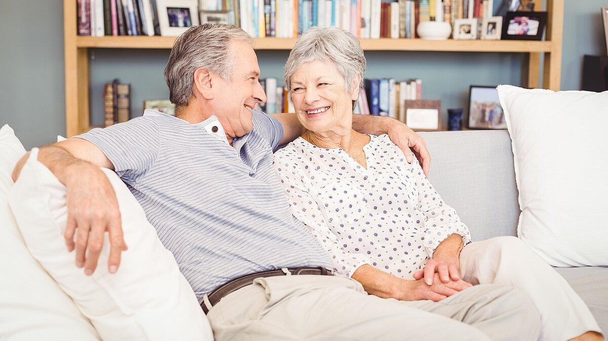 Ein Seniorenehepaar sitzt auf dem Sofa und lächelt sich an. 