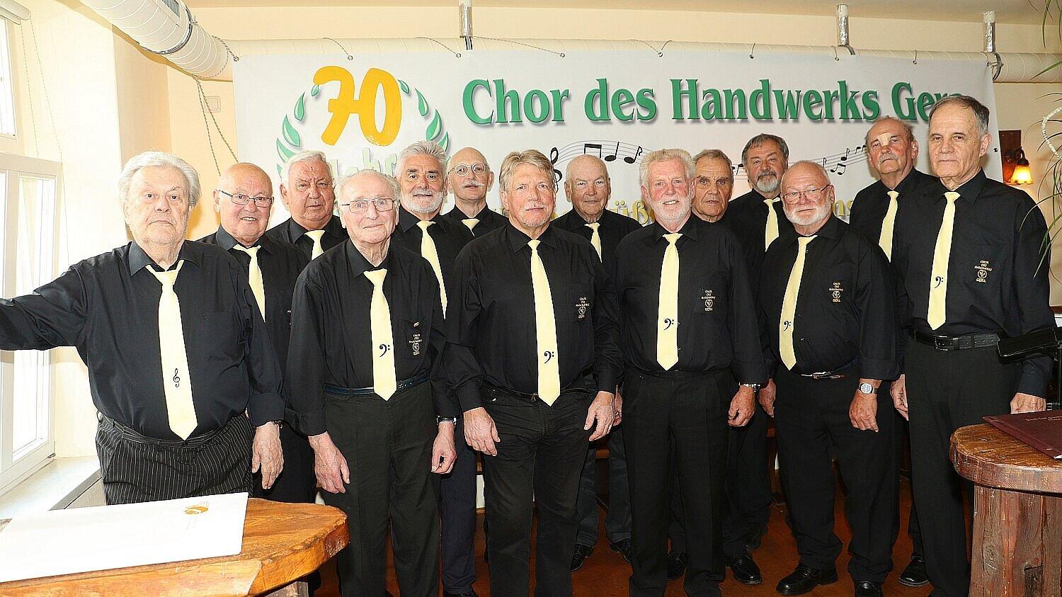 Eine Gruppe an männlich gelesenen Personen höheres Alters in schwarzen Hemden und Blusen stehen vor einen Plakat mit der Aufschrift: "70 Jahre Chor des Handwerks". © Chor des Handwerks Männerchor Stadt Gera e.V.