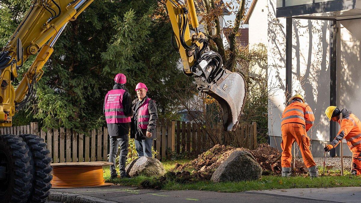 Telekommitarbeiter stehen neben einem Bagger und heben einen Graben aus.. © Telekom