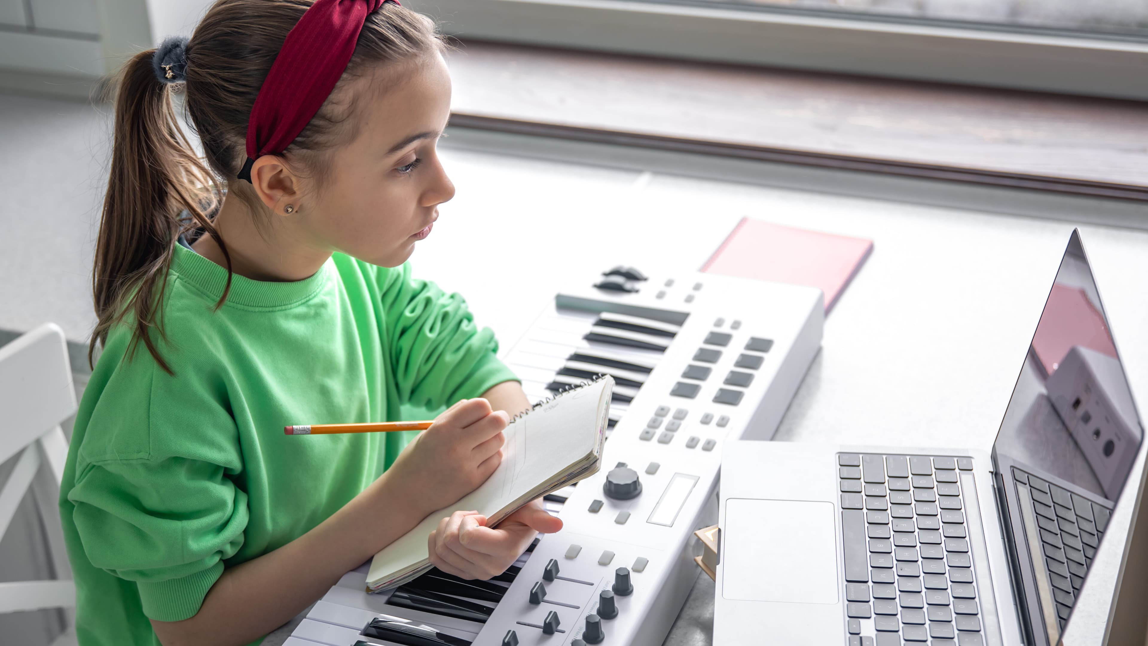 Weiblich gelesenes Kind sitzt mit Keyboard, Stift und Zettel vor Laptop. © Freepik