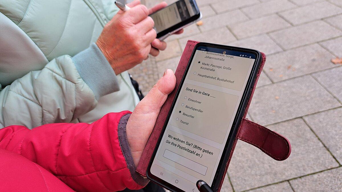 Zwei Frauen halten jeweils ein Smartphone, auf dem ein Ausschnitt der Umfrage zu sehen ist. © Stadt Gera
