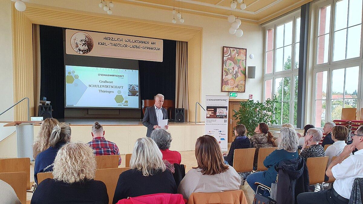 Ein Mann steht in einer Aula vorn und spricht vor einem sitzenden Publikum, welches man von hinten sieht. © IHK Ostthüringen