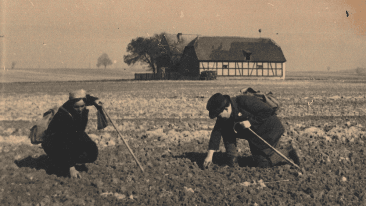 Ehepaar Brause auf Spurensuche auf einem Feld, beide mit Rucksack und Gehstock ausgestattet. Im Hintergrund zwei Häuser und Bäume