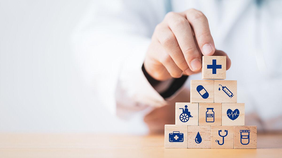 Eine Hand baut Turm aus Würfeln auf. Darauf sind Symbole zur Gesundheit.. © AdobeStock